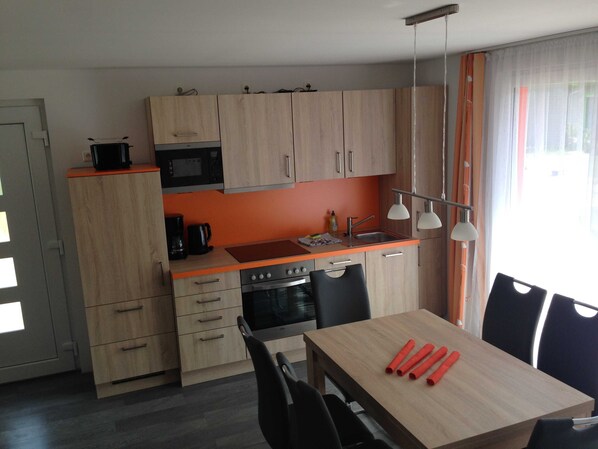 A compact kitchen with a microwave, stove, and sink. There is a dining table with chairs and a window with curtains.
