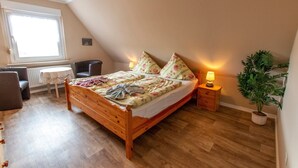 A bedroom with a wooden bed, a small table, a chair, a radiator, a window, a nightstand with a lamp, and a potted plant. - Langfeld, A. Apartment upstairs, 4P - Apartments Langfeld, Christa (Sellin)
