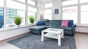 Un salón moderno con un sofá gris, un reposapiés a juego, una mesa de centro blanca y ventanas amplias que ofrecen vistas de un edificio.