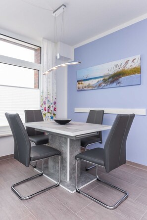 Uma área de refeições com uma mesa e quatro cadeiras, um quadro com uma cena de praia e uma janela com persianas.