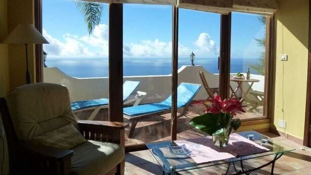 Una habitación con un ventanal grande con vistas a una playa, una silla, una mesa con un jarrón de flores y una lámpara.