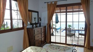 A bedroom with a bed, a dresser, a window with curtains, and a view of an outdoor pool area.