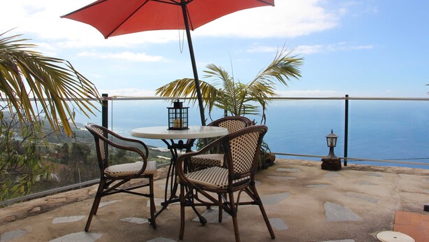 A patio with wicker chairs and a red umbrella, overlooking a scenic view.