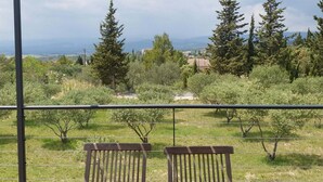 Un balcon avec deux chaises en bois et une rambarde en métal, donnant sur un espace verdoyant avec des arbres et des bâtiments situés au loin.
