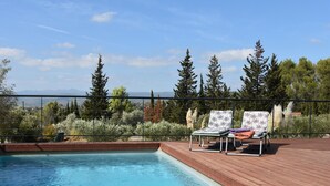 Une piscine avec deux chaises longues sur une terrasse en bois, entourée d’arbres et sous un ciel dégagé.