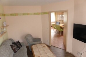 Un salon comprenant un canapé, un fauteuil et un téléviseur. On aperçoit un coin repas à travers une porte ouverte.
