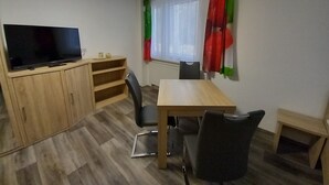 A room with a TV on a wooden cabinet, a table with chairs, a window with curtains, and a radiator.