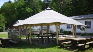 A gazebo with a roof, surrounded by tables and benches, set in a grassy area with trees in the background.