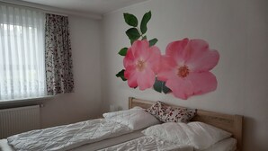 A bedroom with two beds, a radiator, a window with curtains, and a floral wall decoration.