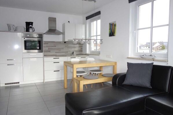A modern kitchen with white cabinetry, a built-in oven, and a dining area with a black leather couch and a wooden table.