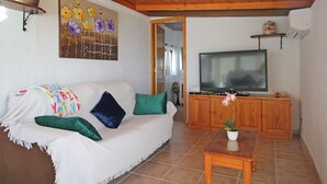 Ein Wohnzimmer mit einem weißen Sofa, grünen Kissen, einem Holztisch, einem Fernseher auf einer Holzkommode und einem Blumenbild an der Wand.
