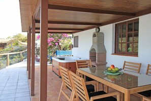 Eine überdachte Terrasse mit einem Glastisch, Holzstühlen, einer Hot Tub und einem Steinofen.