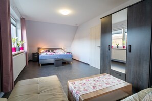 A bedroom with a bed, bedside tables, a desk, a wardrobe, and a window with plants.