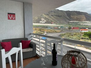 En balkon med en grå sofa, røde puder, et bord med en flaske og glas, og udsigt over et bjergrigt landskab med landbrugsjord.