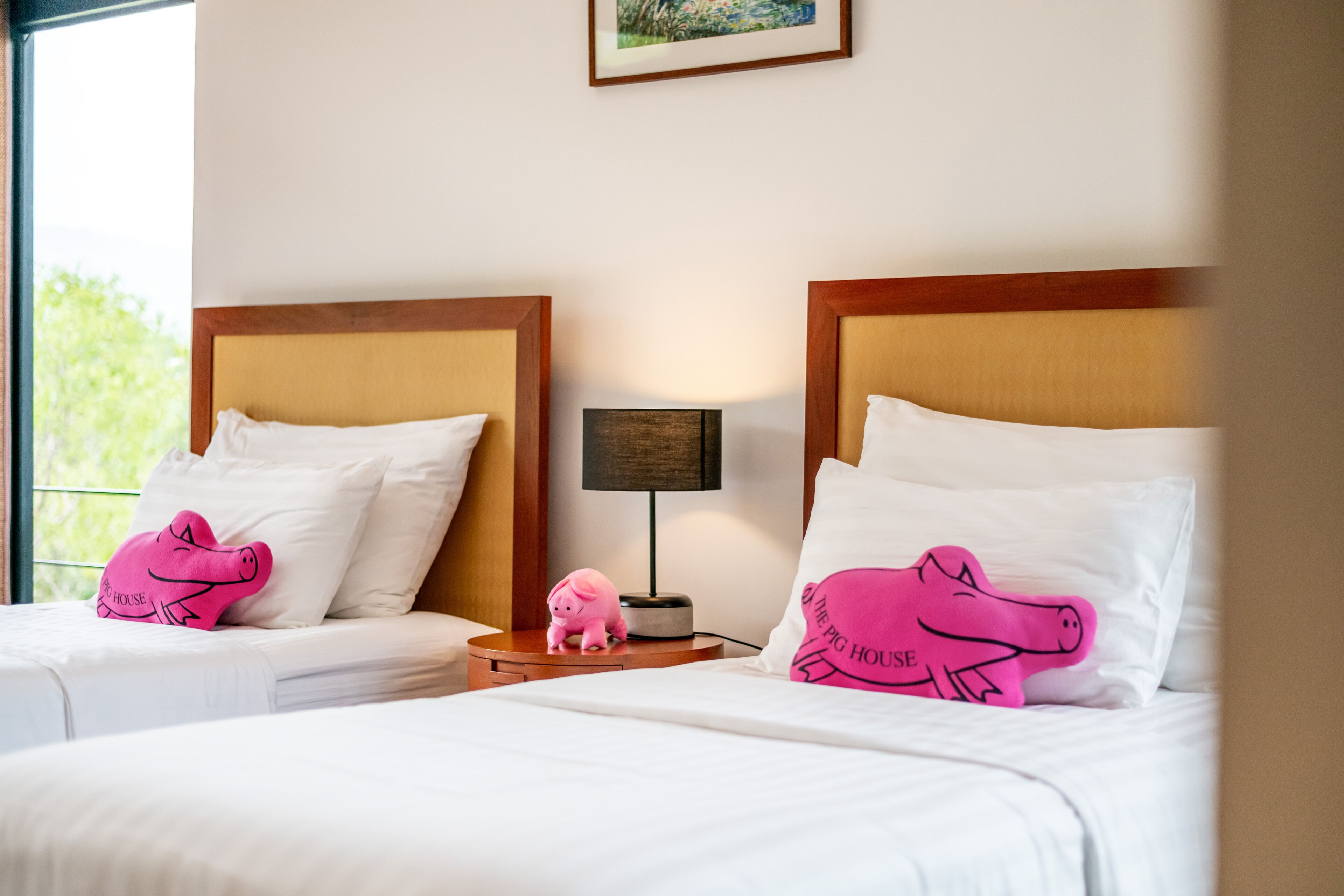 Duas camas com cabeceiras iguais, roupa de cama branca e almofadas em formato de porco rosa.