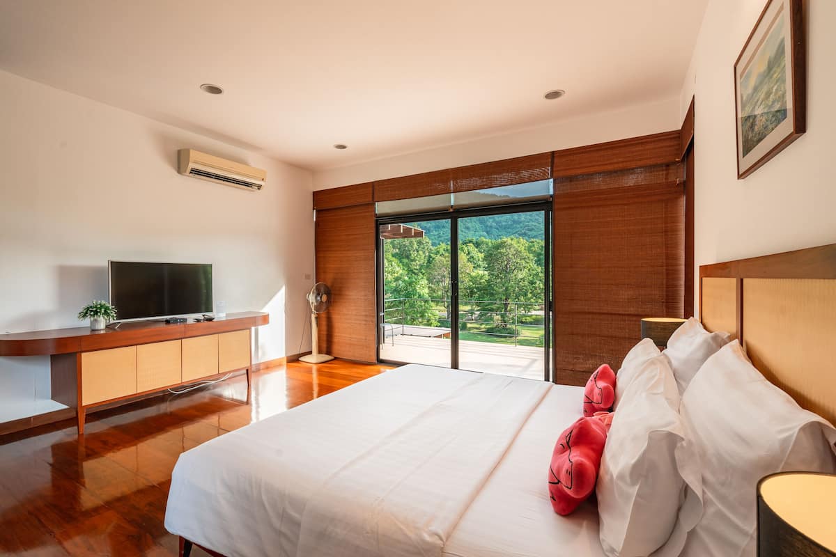 A modern hotel room with a large bed, a flat-screen TV, a wooden desk, and a balcony with a view of greenery.