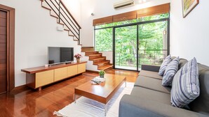 A modern living room with a wooden staircase, a flat-screen TV, a sofa, and large windows offering a view of greenery. - The Pig House Khao Yai (Pak Chong)