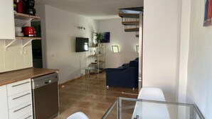 Moderni keittiö, jossa on astianpesukone, kahvinkeitin ja seinälle asennettu televisio. Olohuoneessa on lasipöytä tuoleineen sekä sohva.