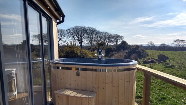 Een houten jacuzzi met treden op een terras, omgeven door een houten leuning en een glazen deur, met uitzicht op een gazon met bomen.