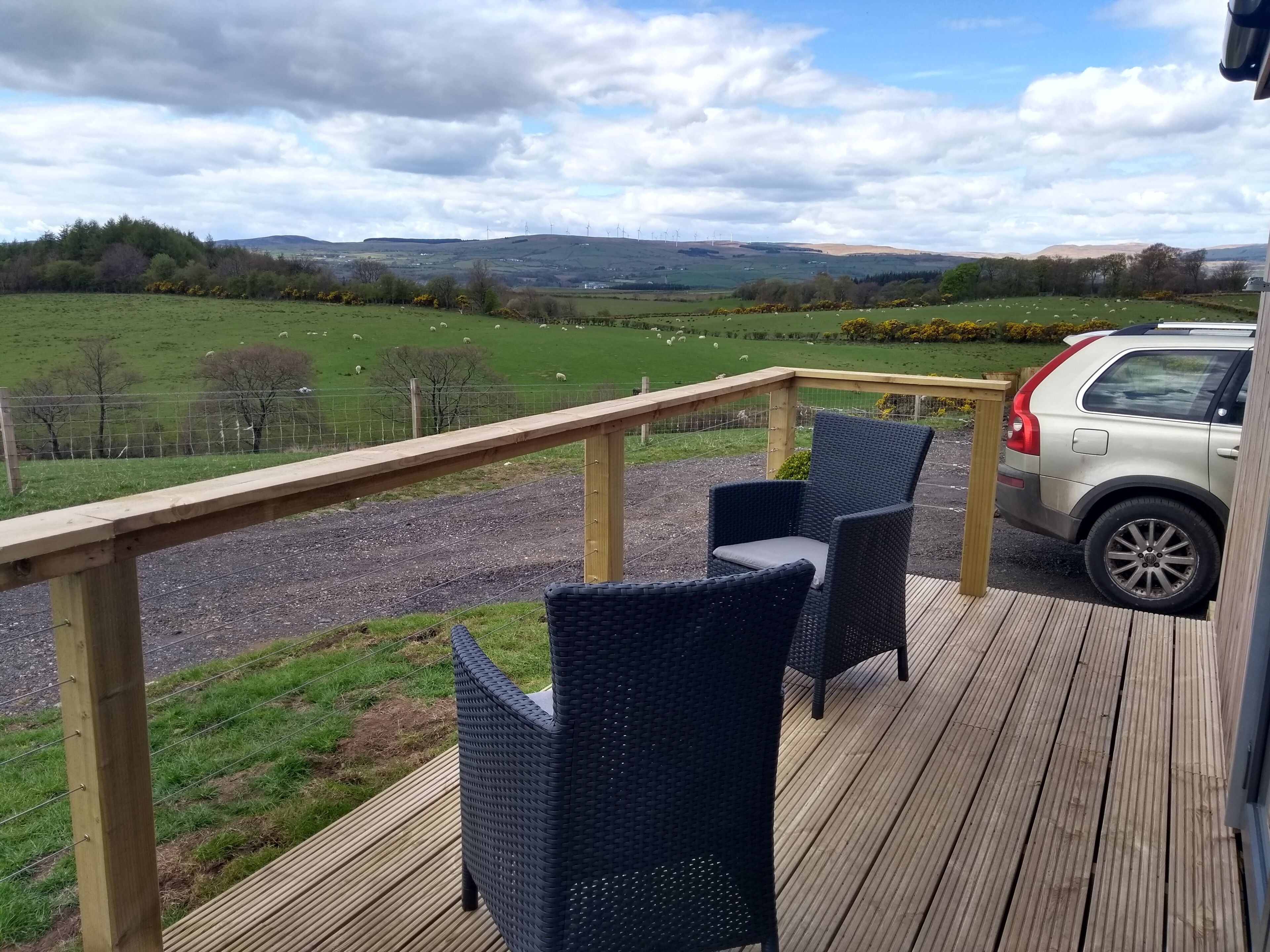 A deck with wicker chairs and a car parked nearby, overlooking a green field with sheep.