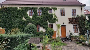 Ein zweistöckiges Haus mit rotem Ziegeldach, efeuüberwucherten Mauern und einem kleinen Garten mit Kübelpflanzen und einer hölzernen Struktur.