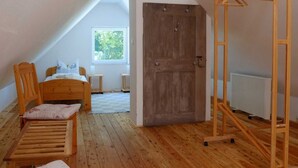 Ein Zimmer mit schrägem Dach, einem hölzernen Bett, einem Holzstuhl, einem Wäscheständer aus Holz und einem Fenster mit Blick ins Grüne.