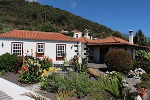 A white house with a red-tiled roof, surrounded by a garden with various plants and flowers.