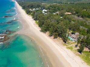 An aerial view of a beach with clear turquoise waters, a sandy shore, and a cluster of houses surrounded by dense greenery.