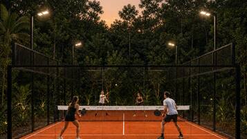 An outdoor tennis court with players, surrounded by trees and illuminated by street lamps.
