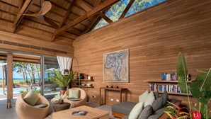 A modern living room with a wooden ceiling, a large window overlooking a pool, and a bookshelf filled with books. - The Pines (Takua Thung)