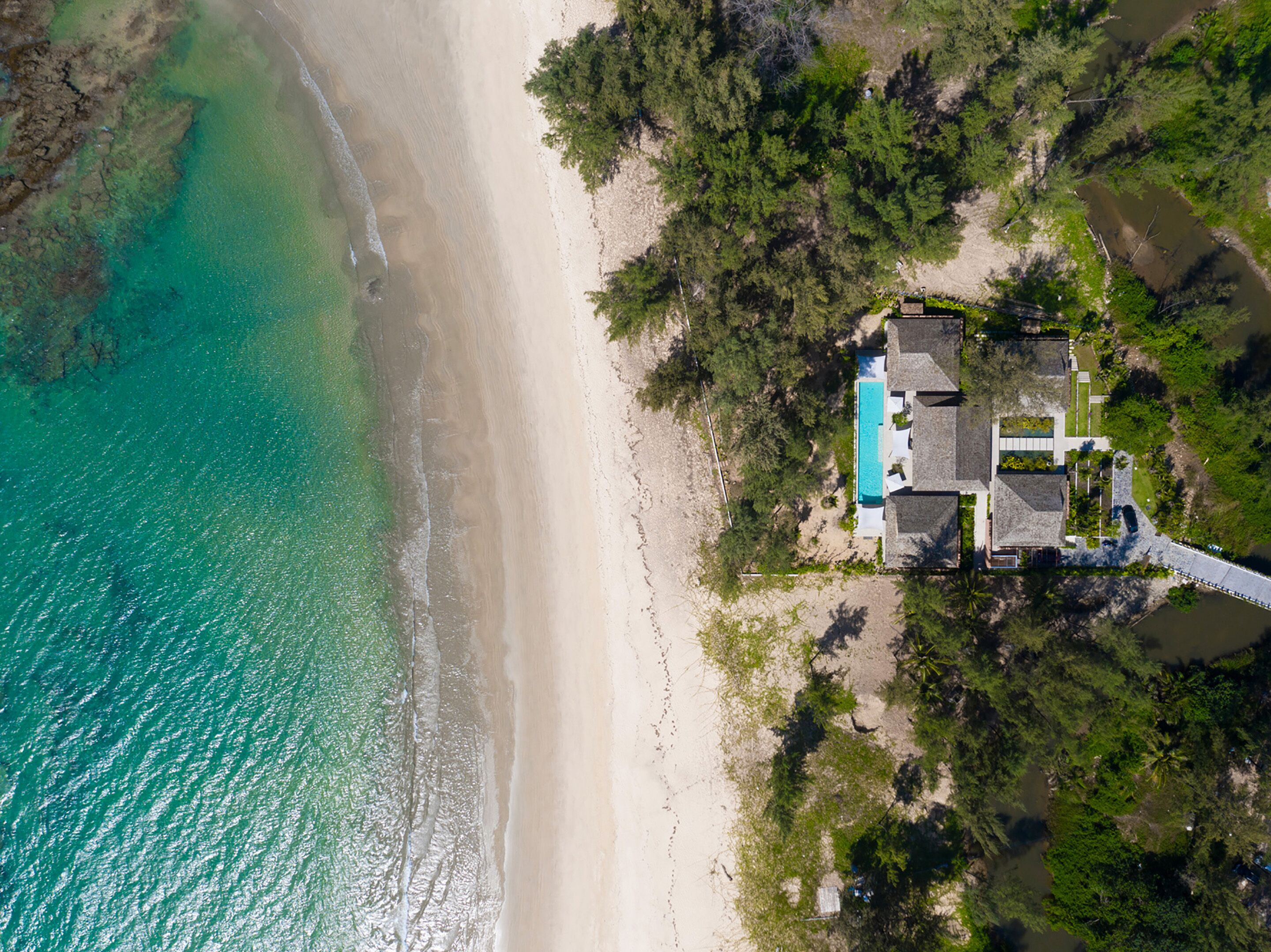 An aerial view of a coastal property with a swimming pool, beach access, and dense vegetation.
