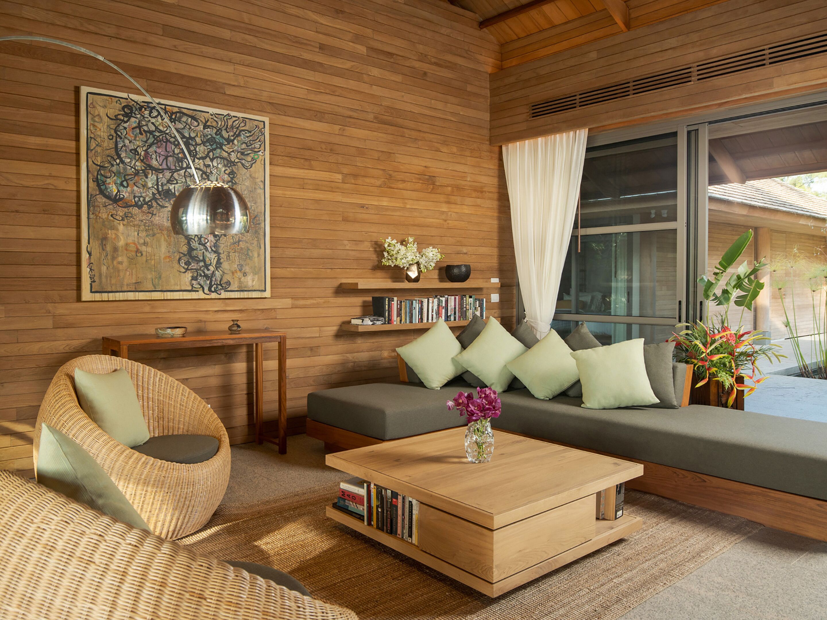 A modern living room with a wooden floor, a sofa with cushions, a coffee table, a wicker chair, a bookshelf, and a large abstract painting.