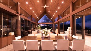 A spacious dining area with a high ceiling and wooden beams, featuring a large table surrounded by white chairs, a bar area, and a view of the night sky. - Suralai by Elite Havens (Koh Samui)