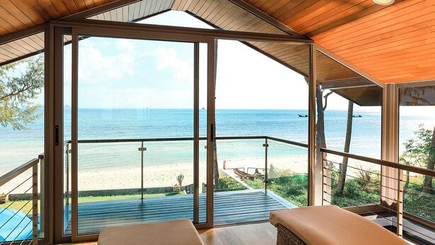 Una habitación con un amplio ventanal que da a un balcón con vistas a la playa. La habitación tiene techo de madera y una cama con colchón color beige.