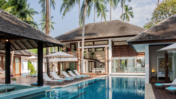 A pool area with lounge chairs and umbrellas, surrounded by palm trees and a modern building with large windows. - Ban Suriya (Koh Samui)