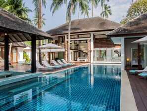 A pool area with lounge chairs and umbrellas, surrounded by palm trees and a modern building with large windows. - Ban Suriya (Koh Samui)