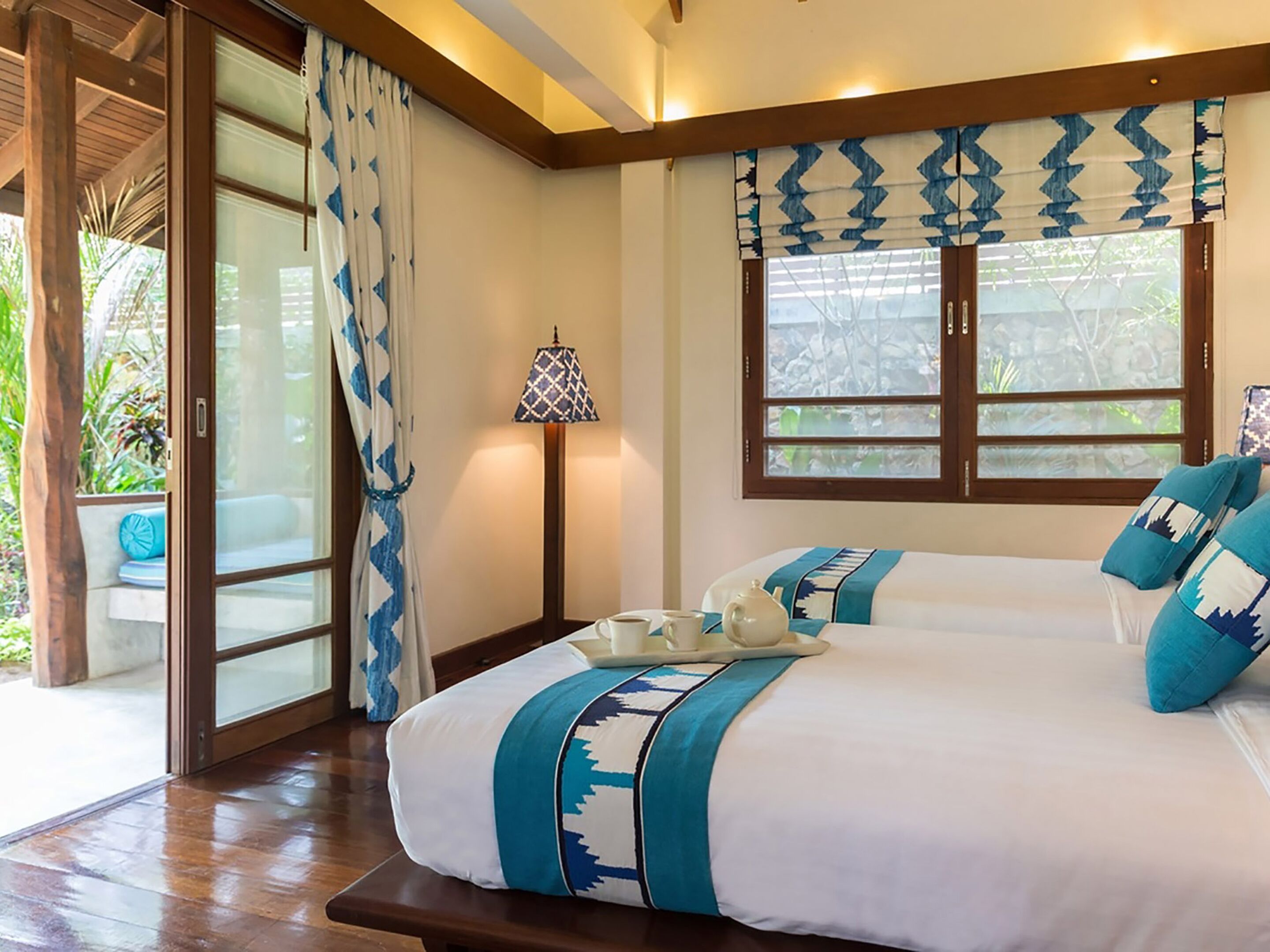 A hotel room with a large bed, a bedside table with a lamp, and a window with a view of greenery.