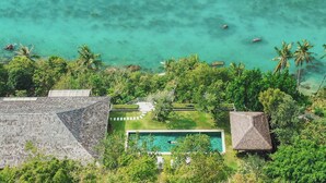 An aerial view of a resort with a swimming pool surrounded by lush greenery and palm trees, overlooking clear turquoise waters. - Cape Laem Sor Estate (Koh Samui)