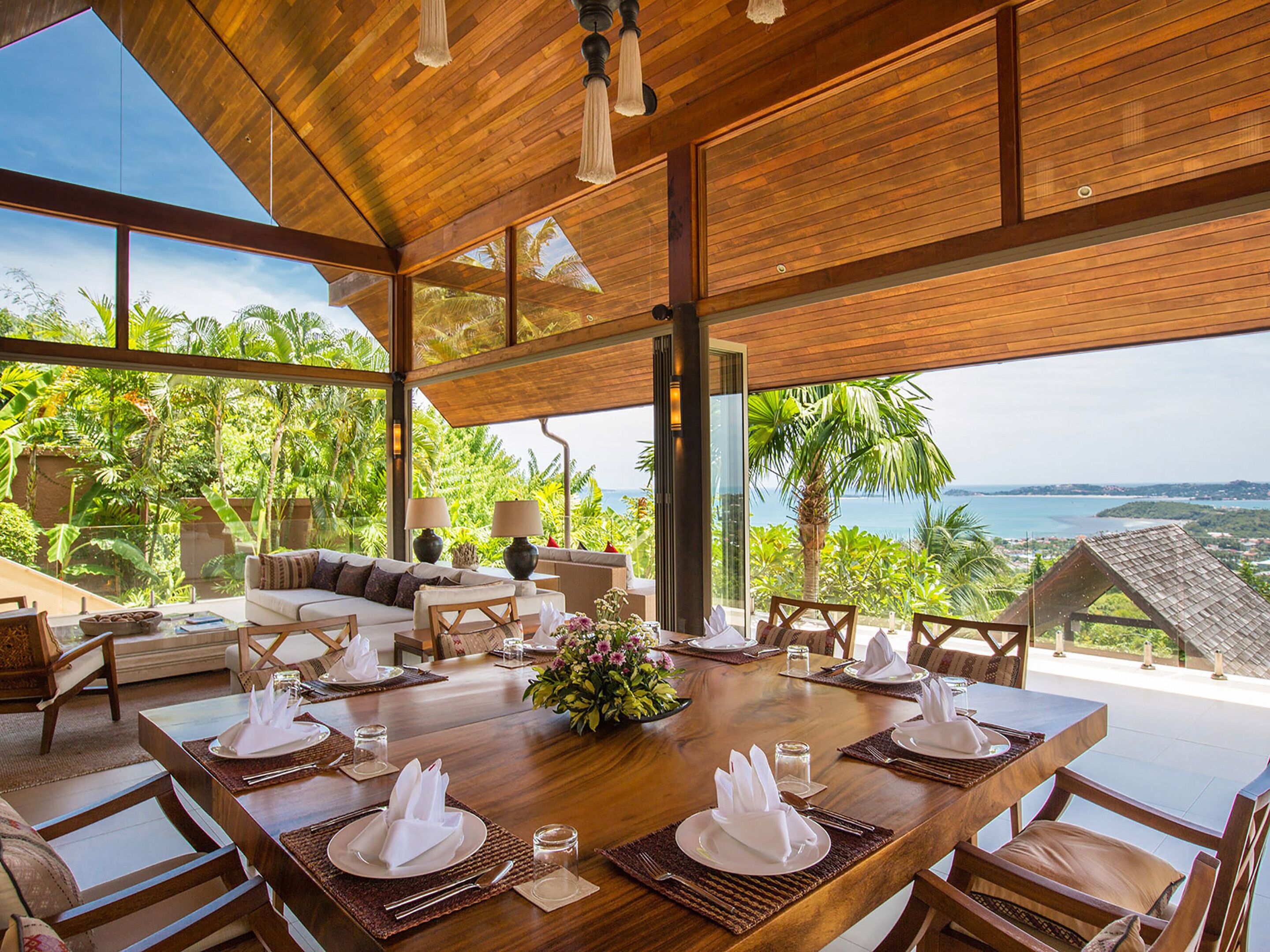 Un comedor amplio con una mesa de madera dispuesta para seis personas, con vistas a un jardín exuberante y una vista panorámica del océano.