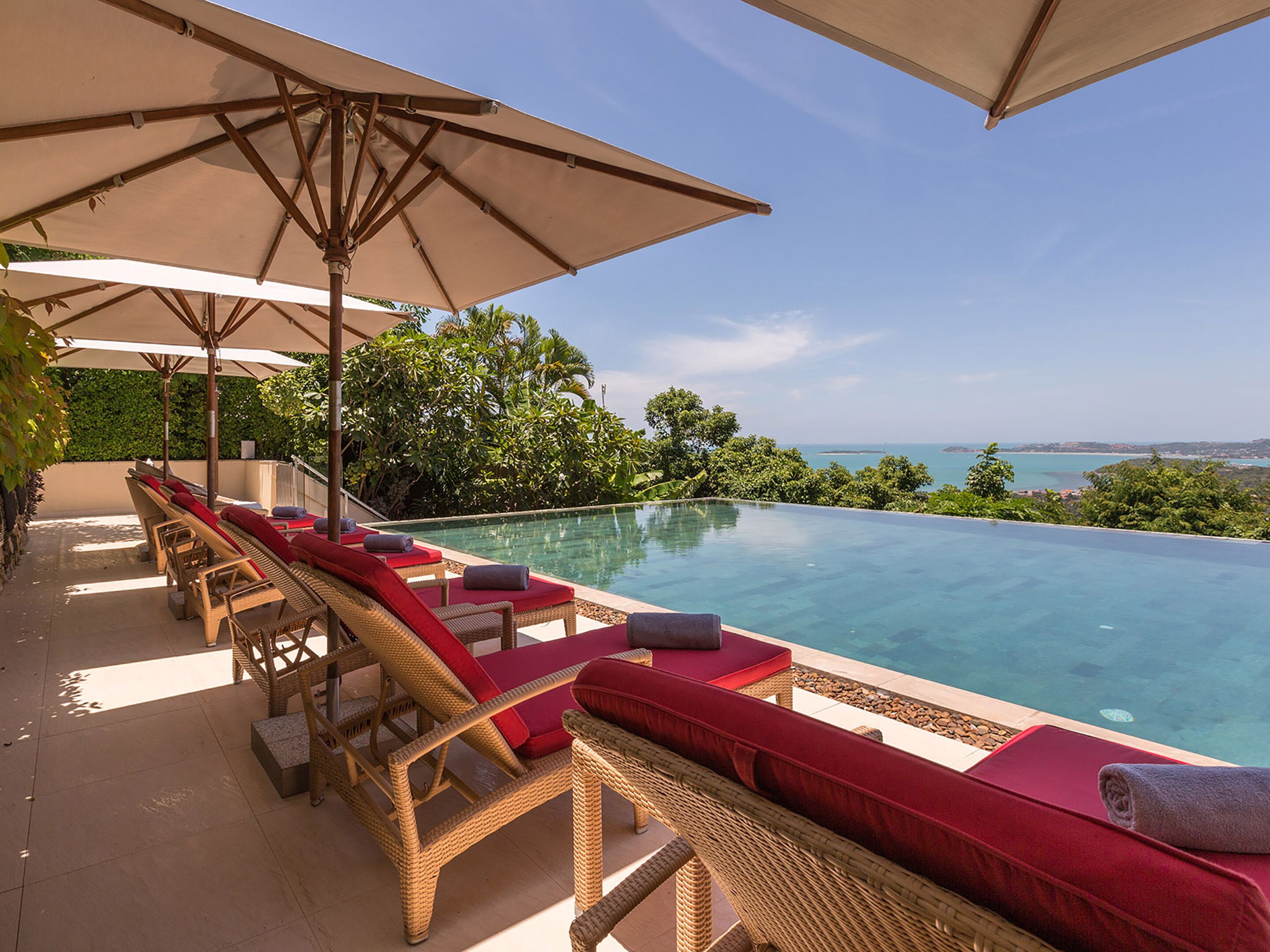 An infinity pool with wicker lounge chairs and red cushions, surrounded by lush greenery and a clear view of the sea.