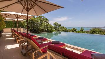 An infinity pool with wicker lounge chairs and red cushions, surrounded by lush greenery and a clear view of the sea.