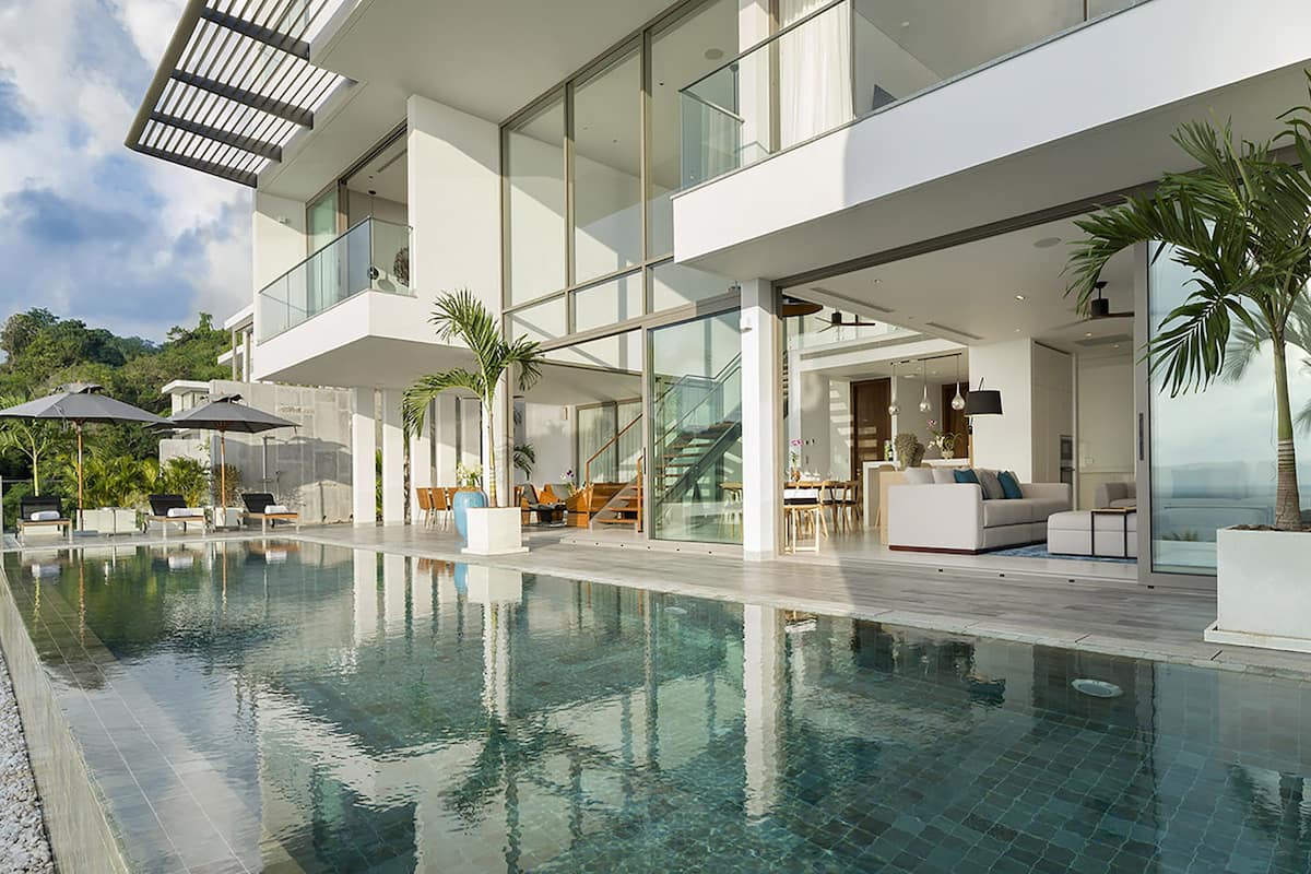 A modern poolside area with a glass-enclosed infinity pool, outdoor seating, and a staircase leading to an upper level.