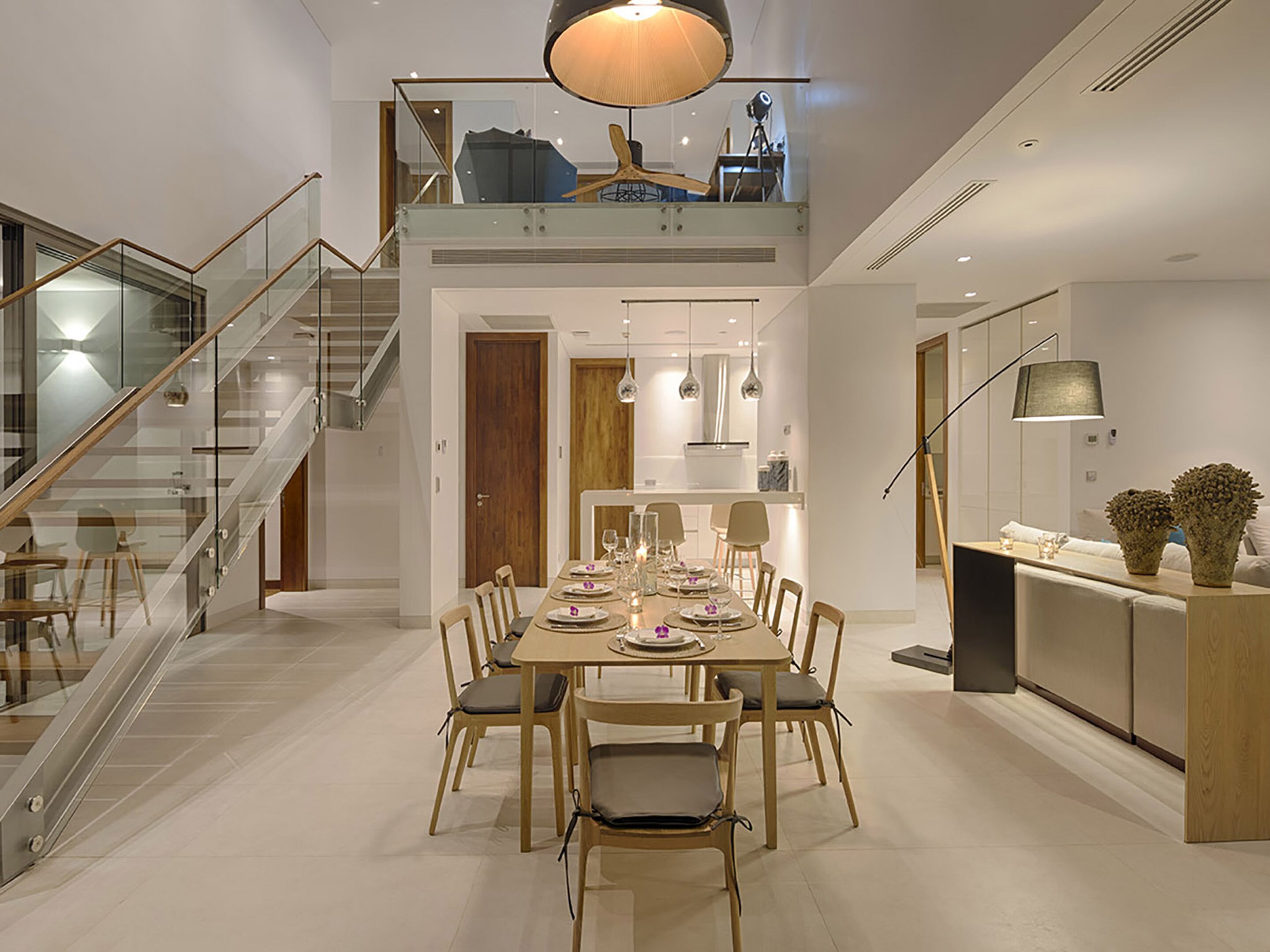 Een moderne eetruimte met een lange tafel voor zes personen, een trap met glazen leuningen en een keuken met hanglampen.