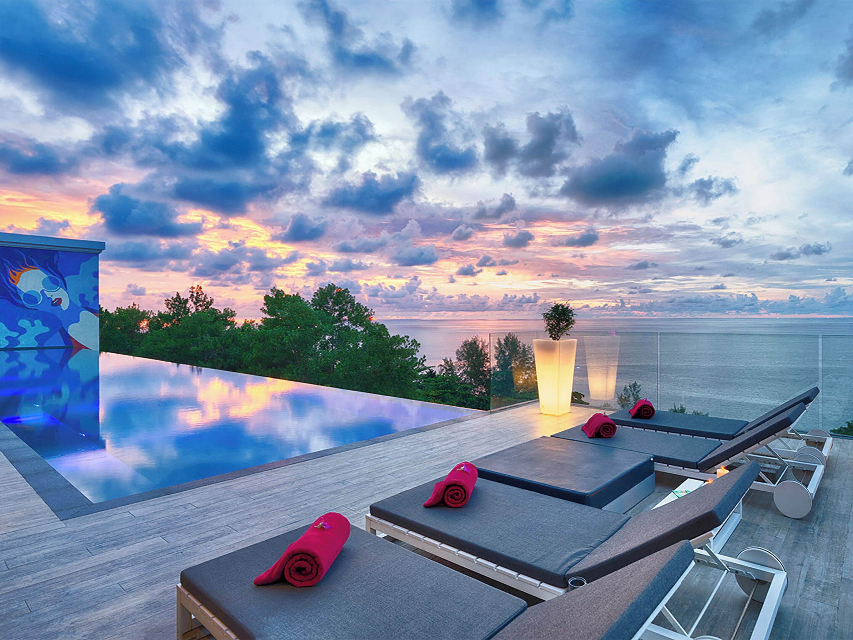 An infinity pool with lounge chairs and a potted plant, overlooking a body of water with a sunset sky.