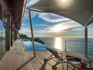 Una terraza de madera con una piscina infinita de cristal, sillas de descanso y vista al océano al atardecer.