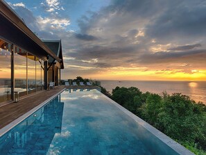 Una piscina infinita moderna con terraza de madera, con vistas a un paisaje marino al atardecer.