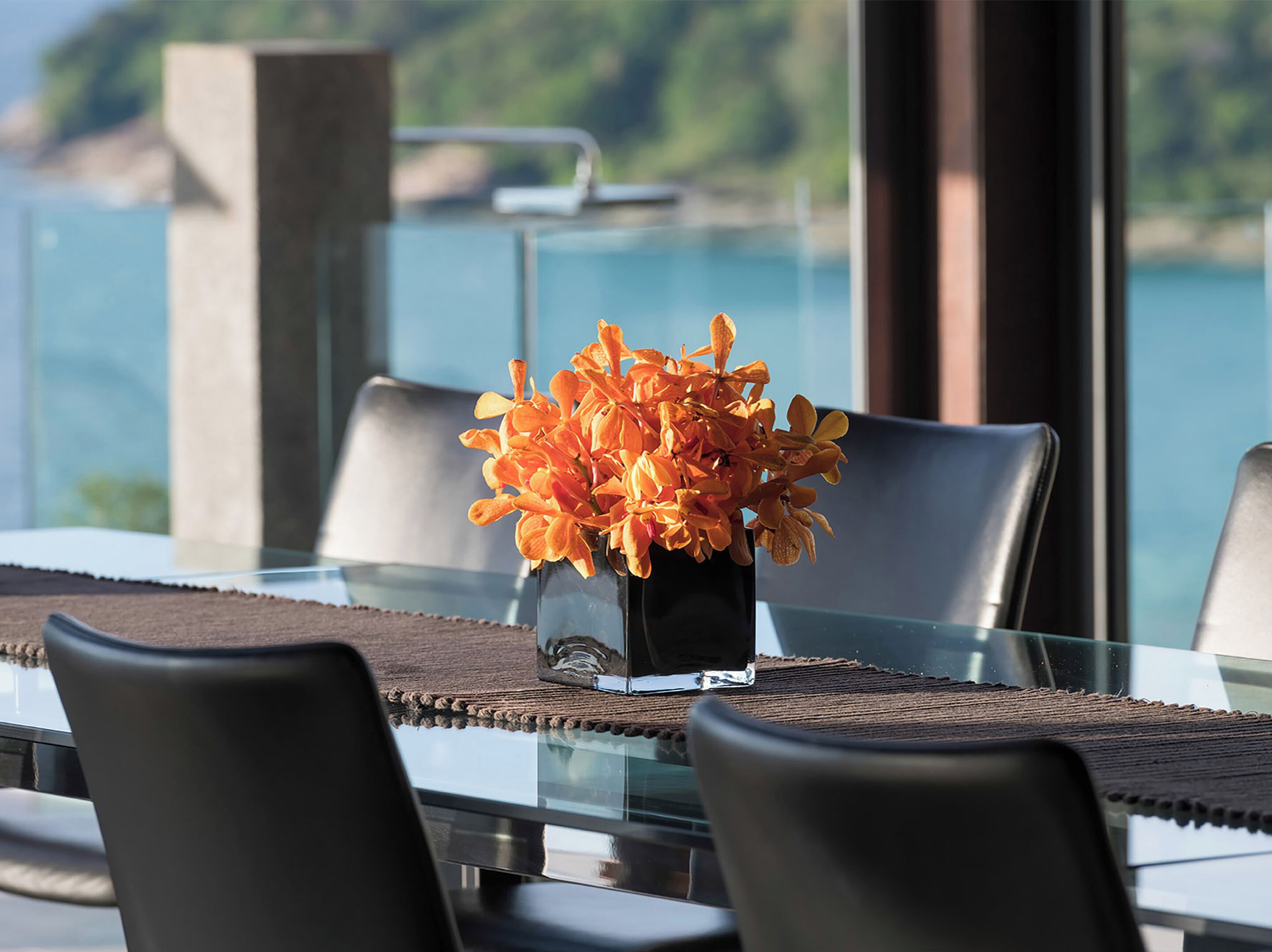 Ein Esstisch mit Stühlen, eine Vase mit orangefarbenen Blumen und Meerblick durch große Fenster.