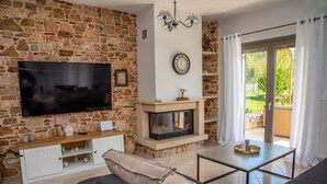 A living room with a stone wall, a flat-screen TV, a fireplace, a sofa, and a sliding glass door to the outdoors. - Giannakis Villa (Kerkyra)