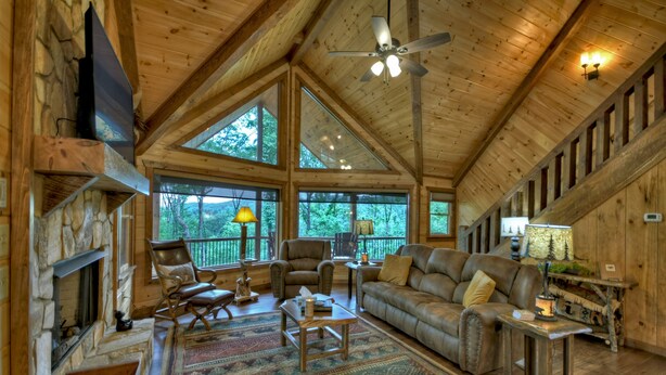 A spacious living room with a stone fireplace, leather furniture, and large windows offering a view of the outdoors. - Deer Trails Cabin - Blue Ridge (Cherry Log)
