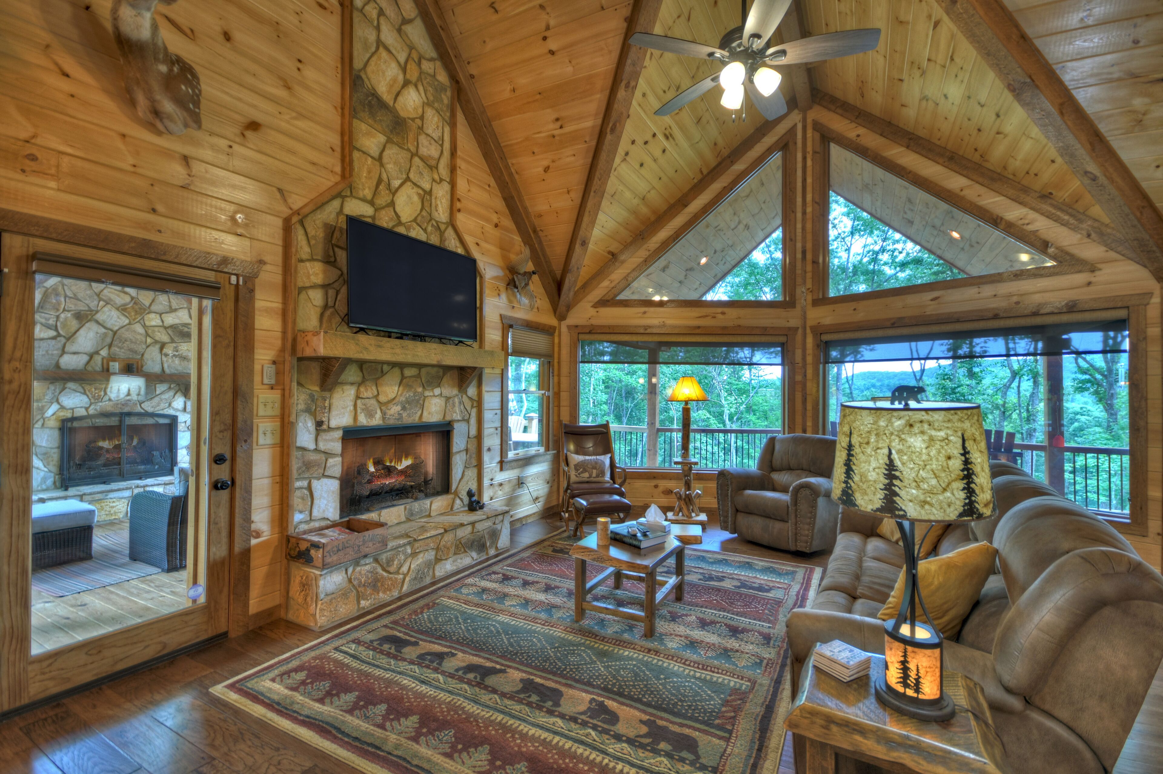 A spacious living room with a stone fireplace, wooden ceiling, and large windows offering a view of the outdoors.